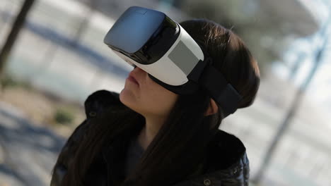 Serious-young-dark-haired-woman-in-warm-black-waistcoat-putting-on-virtual-reality-glasses-in-early-autumn-park